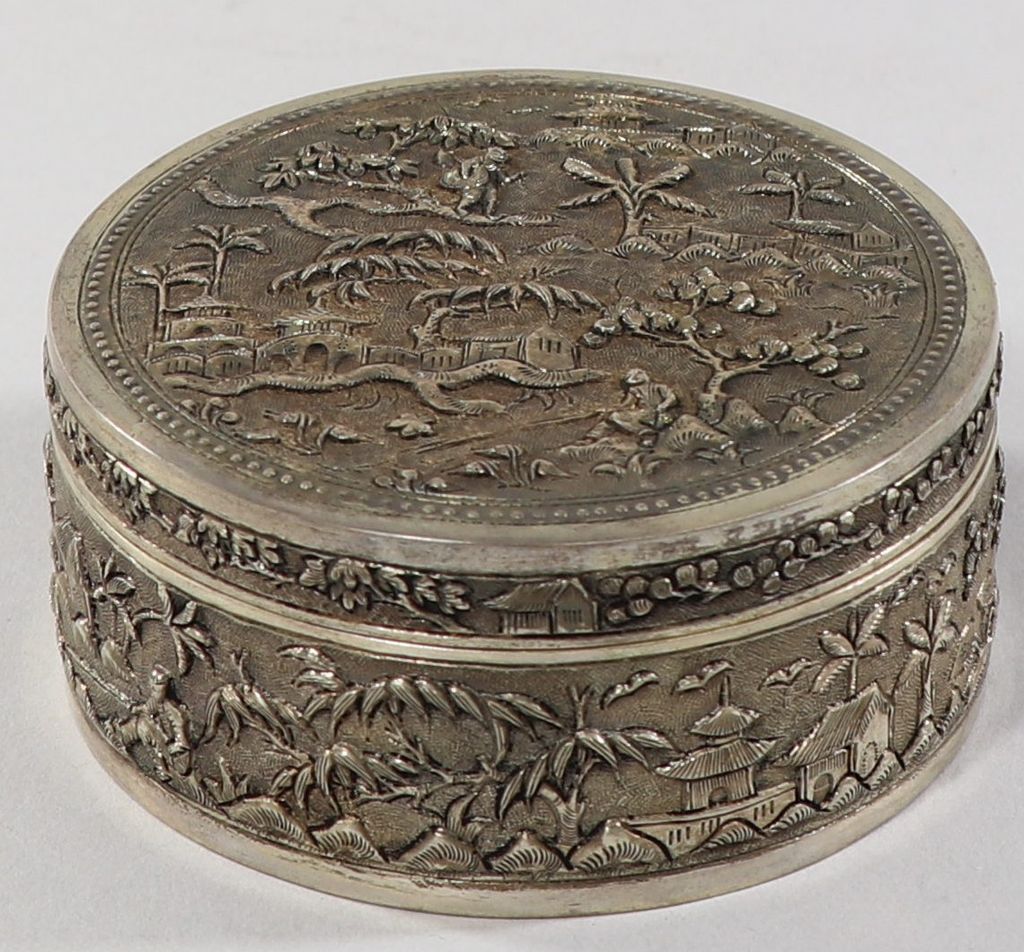 ກັັບໃສ່ແປ້ງ ສະແຕນເລດ ໂອບດ້ວຍເງິນ ດ້ານຫຼັງມີຮູບເຮືອ, ຕົ້ນໄມ້ ແລະ ຄົນ.