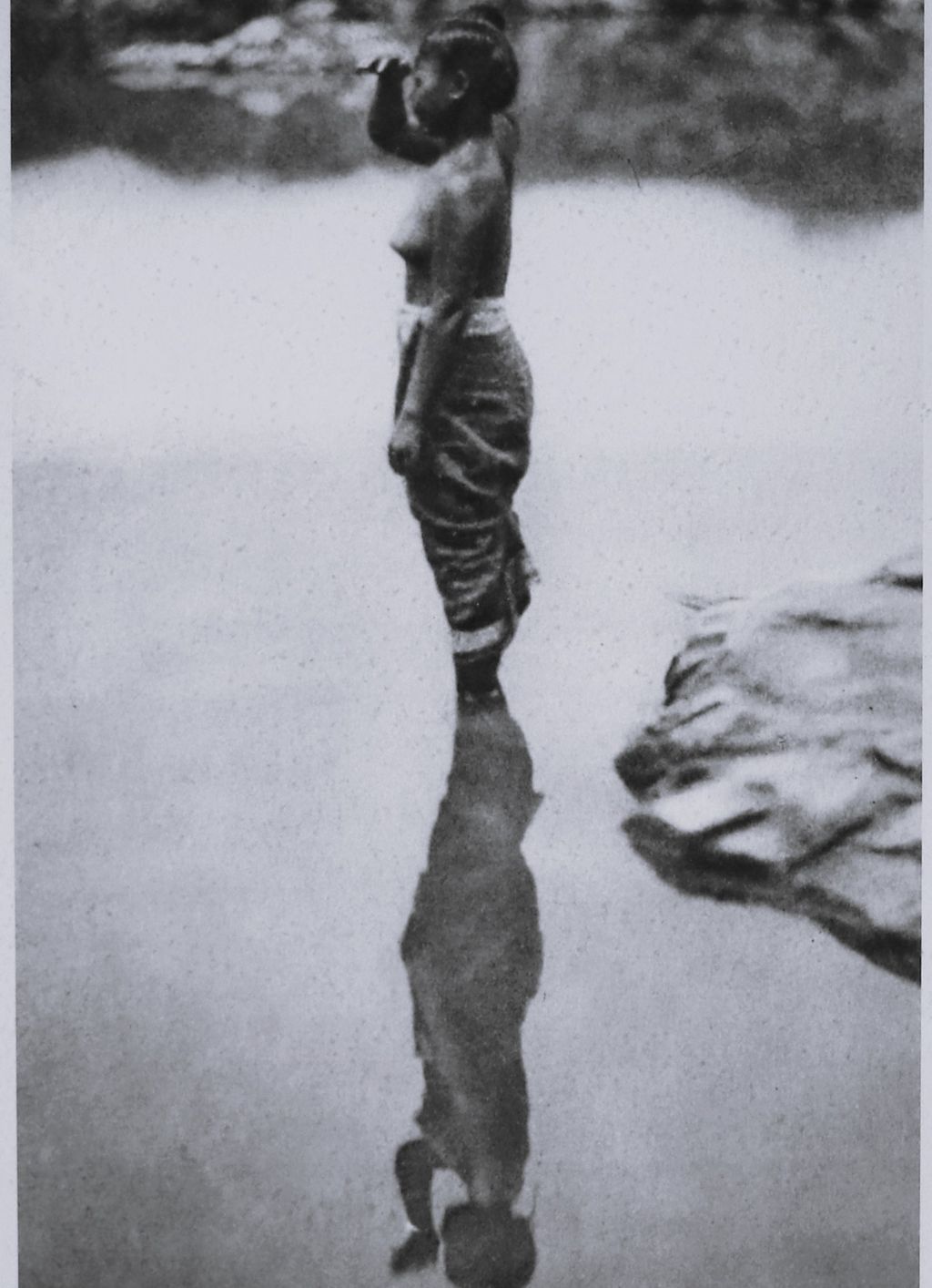 ຮູບ​ພ​າບຂາວດຳ ແມ່ຍິງລາວຄົນໜຶ່ງກຳລັງຍືນຢູ່ແຄມນໍ້າຈ້ອງເບິ່ງເຮືອແລ່ນຜ່ານ ປີ 1930